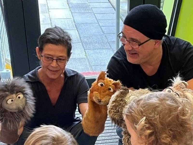 Zwei SchauspielerInnen spielen mit Handpuppen das Handpuppenspiel "Der Wandertag": Links eine Frau mit einem Igel und einem Eichhörnchen, rechts ein Mann mit einem Hasen und einer Eule.